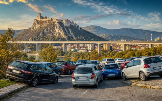 Ilustračný pohľad na Považský hrad, diaľničný viadukt a Považskú Bystricu s autami zaparkovanými nadivoko v popredí.