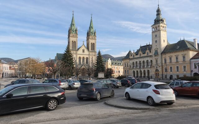 Ilustračný pohľad na Kostol svätého Ondreja a radnicu v Ružomberku s nepravidelne zaparkovanými autami v popredí.
