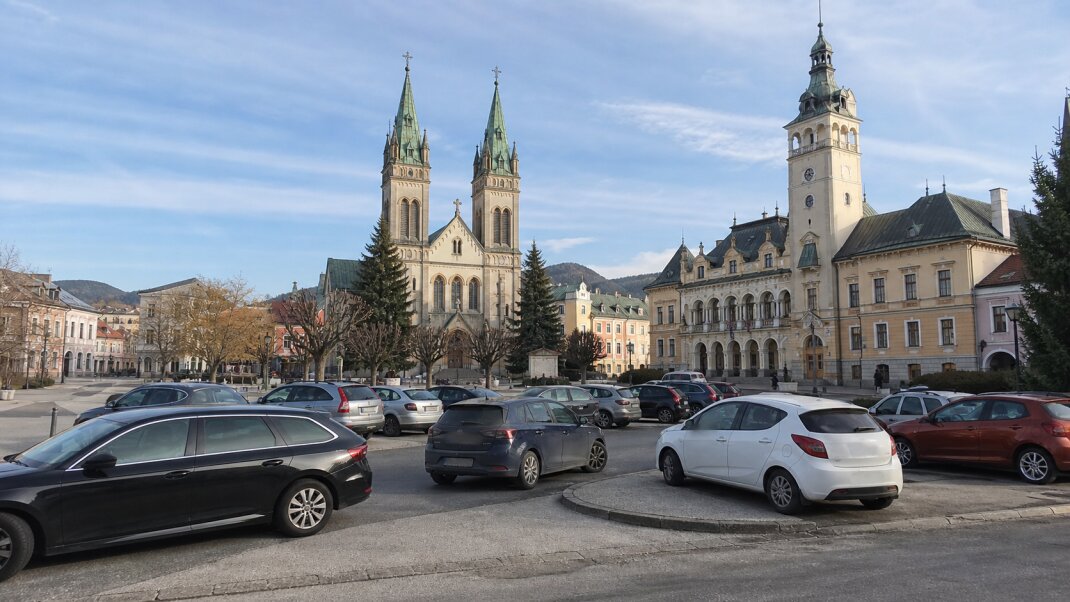 Ilustračný pohľad na Kostol svätého Ondreja a radnicu v Ružomberku s nepravidelne zaparkovanými autami v popredí.