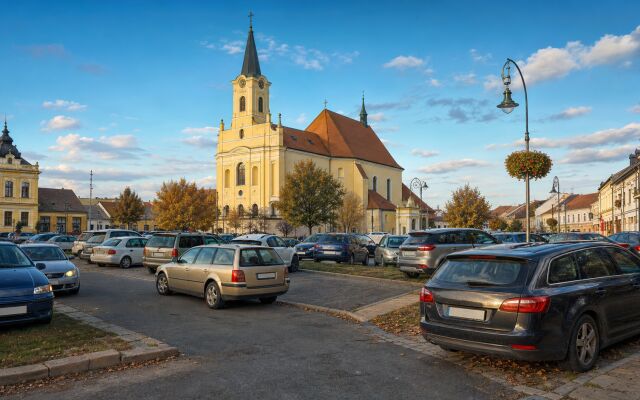 Ilustračný pohľad na Kostol svätého Juraja a centrum Dunajskej Stredy s chaoticky zaparkovanými autami v popredí.