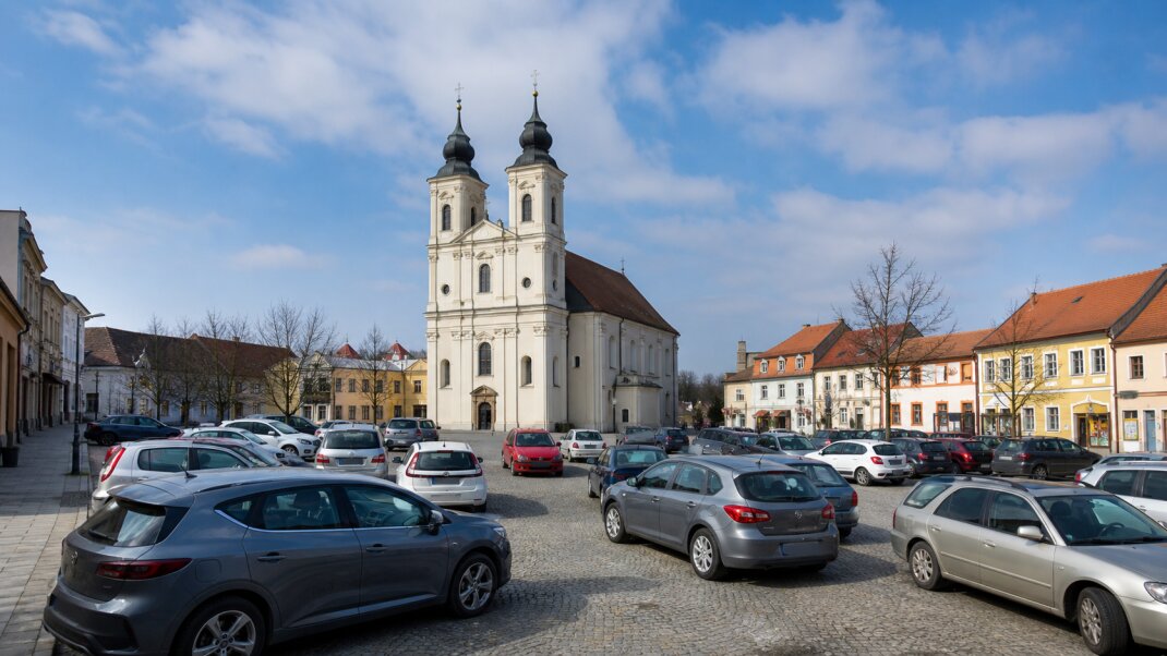 Ilustračný pohľad na historický kostol a centrum Senice s autami zaparkovanými nadivoko v popredí.